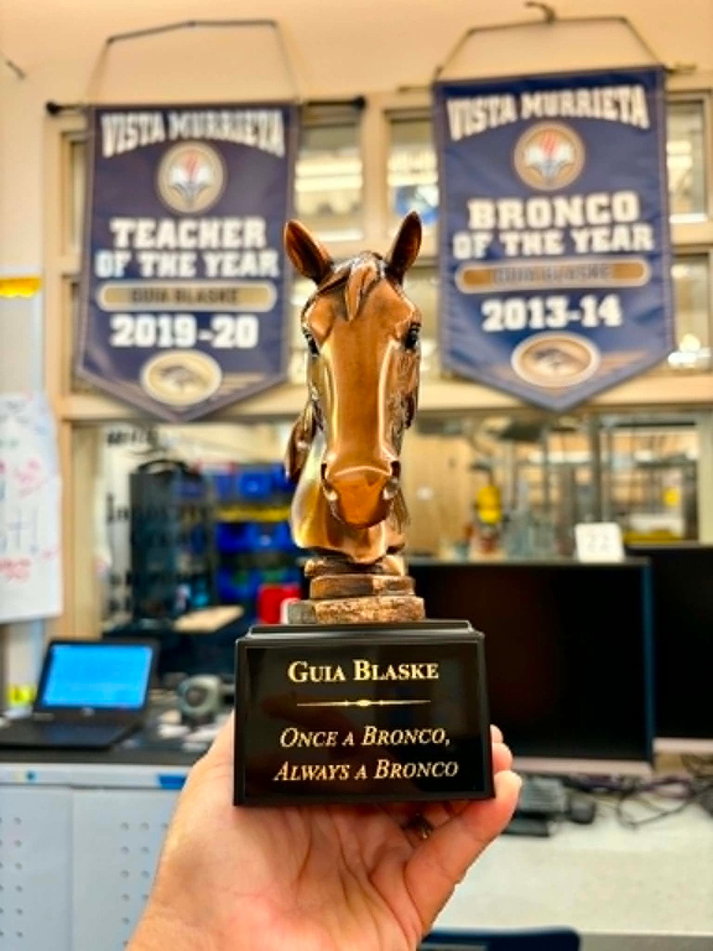 Bronze horse trophy engraved Guia Blaske, Once a Bronco, Always a Bronco, held up in front of Vista Murrieta Teacher of the Year 2019-20 and Bronco of the Year 2013-14 banners